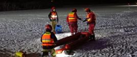 Zdjęcie przedstawia strażaków podczas nocnych ćwiczeń lodowych.
W tle samochody strażackie, zmarznięte jezioro.
