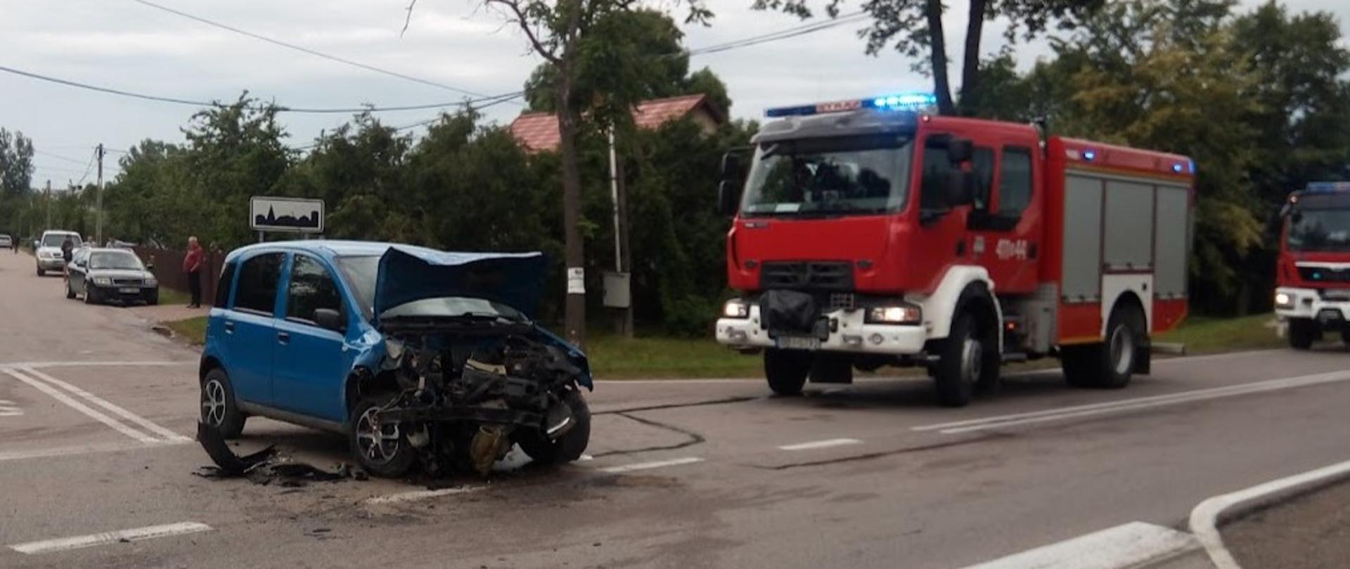 Uszkodzony samochód osobowy stojący na jezdni po zderzeniu z busem. W aucie całkowicie zniszczony przód. W tle samochód ratowniczo-gaśniczy.