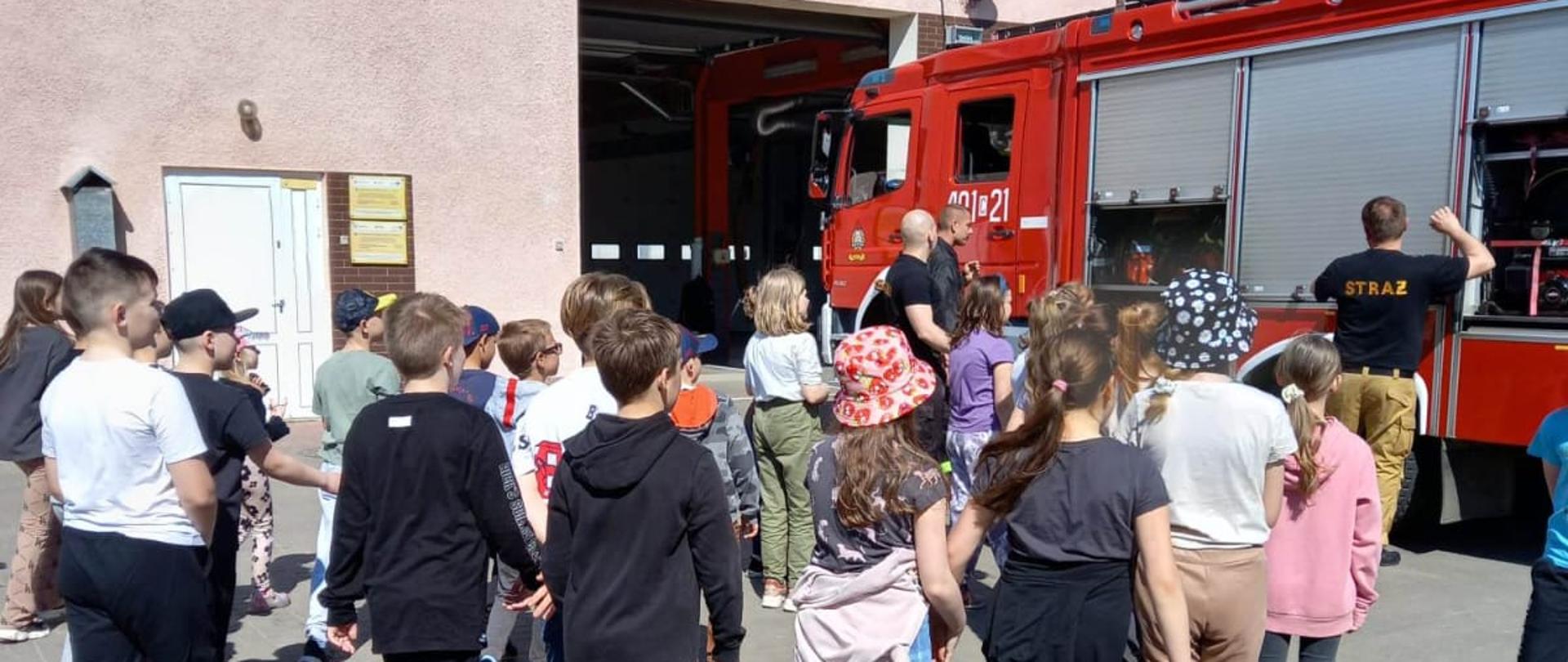 Strażacy wraz z dziećmi na tle budynku JRG oraz samochodu pożarniczego opowiadający na temat pracy w PSP