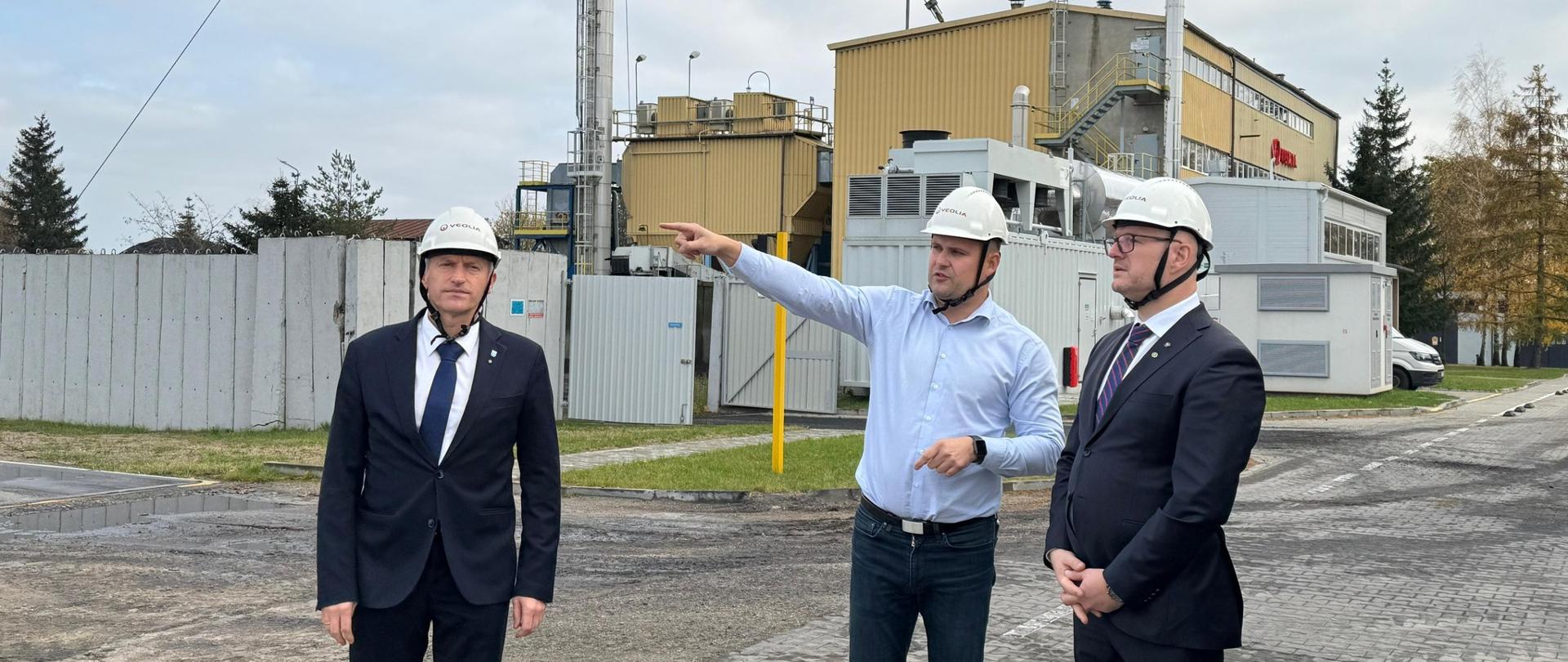 Trzech mężczyzn w garniturach i kaskach stoi przed elektrownią. 