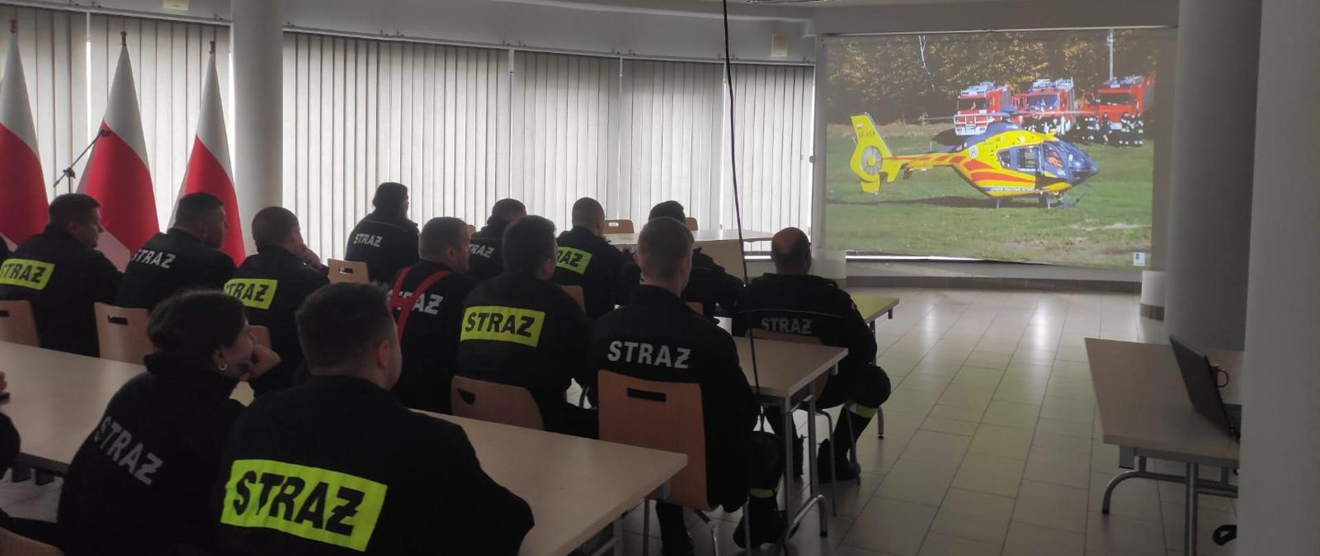 Kilkudziesięciu mężczyzn ubranych w czarne ubrania koszarowe strażackie z odblaskowymi paskami, czarnym butami, siedzą w ławkach. W tle wyświetlana jest prezentacja, żaluzje koloru szarego