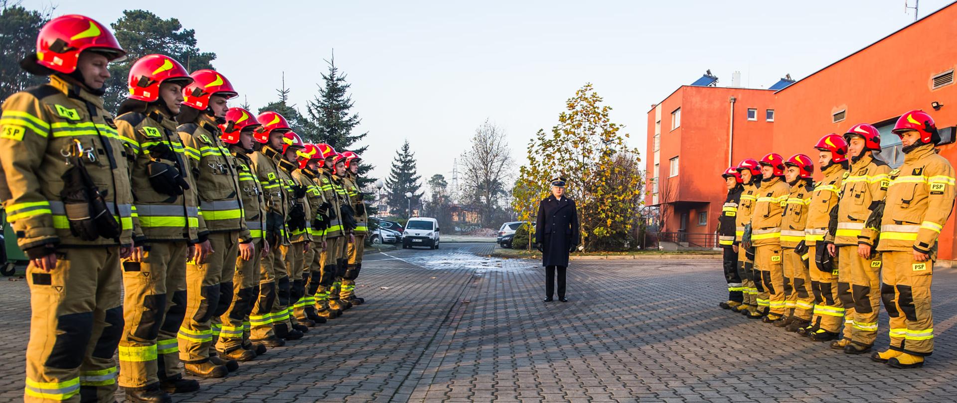 Na zdjęciu funkcjonariusze zmiany służbowej, umundurowani w odzieży specjalnej podczas uroczystej zmiany służby z okazji święta odzyskania niepodległości. Na drugim tle budynek KP PSP Otwock 