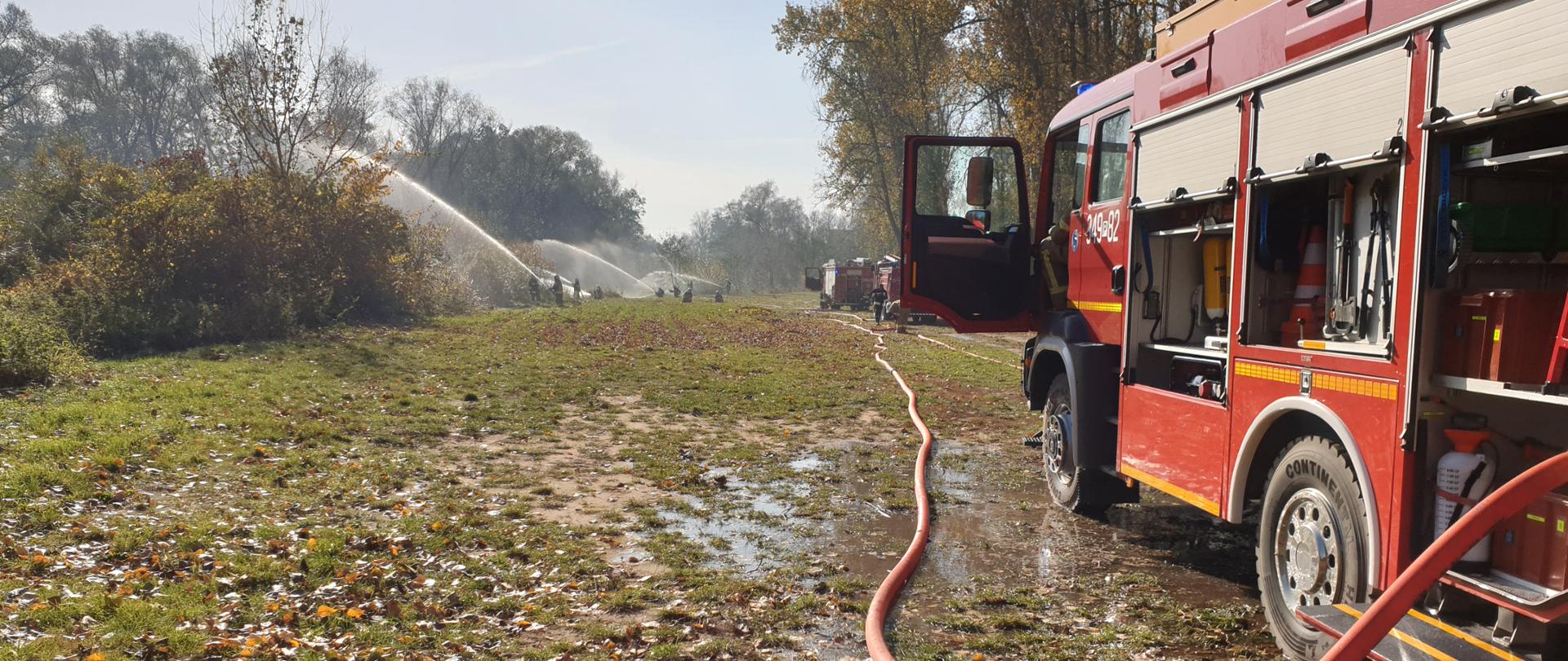 Po prawej stronie samochód pożarniczy. Otwarte żaluzje boczne. Od auta poprowadzona jest linia zasilająca. Wąż ma kolor czerwony. W tle widać strażaków, którzy podają prądy wody obok nich samochody pożarnicze. Niebo lekko zachmurzone, pogoda słoneczna. Po prawej linia drzew, po lewej zarośla.