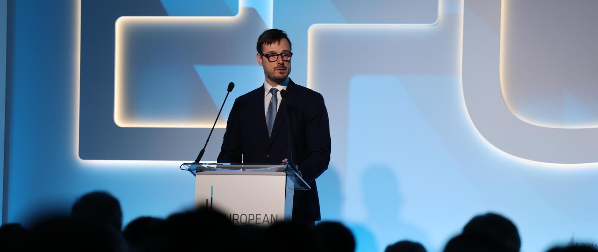 Minister Jakub Jaworowski podczas EFC 2024. Minister stoi przy mównicy, przed nim publiczność zwrócona tyłem do aparatu.