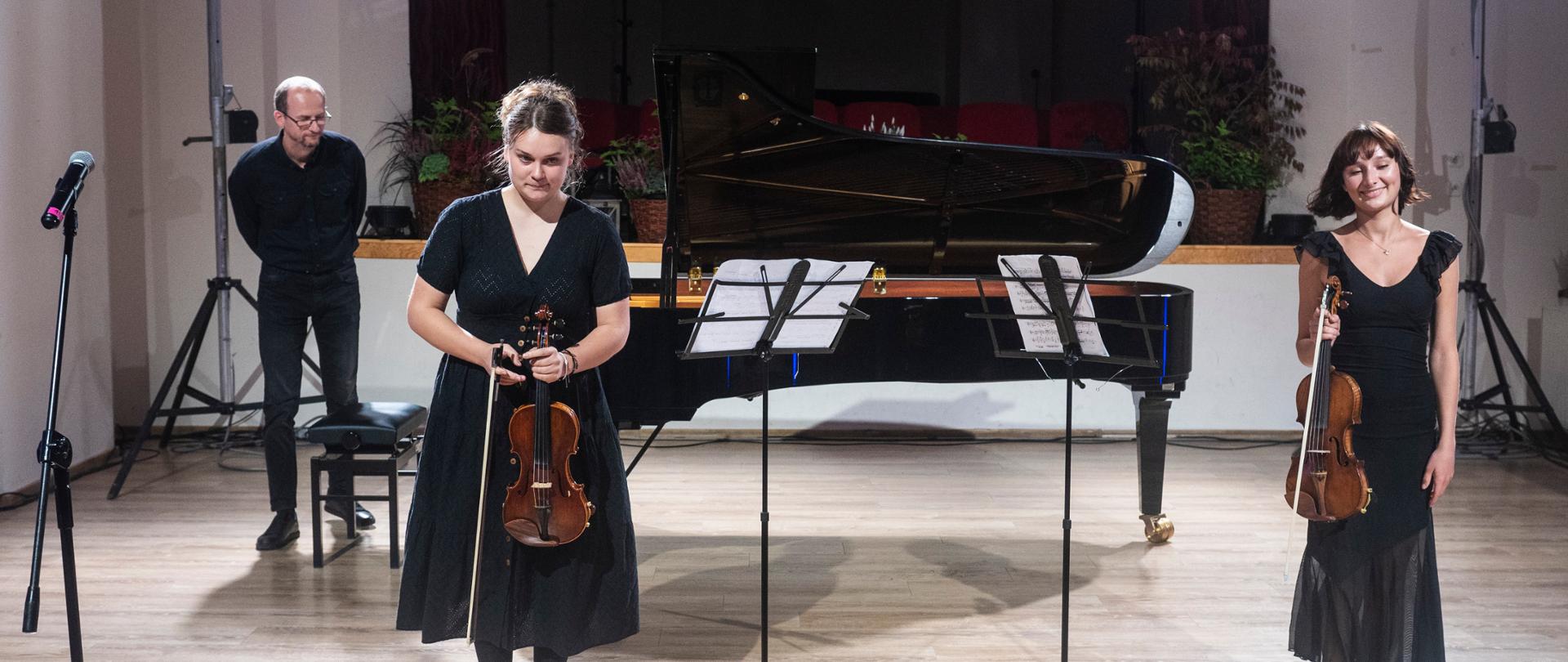 Na scenie dwie skrzypaczki przed występem, trzymają pionowo w rękach skrzypce i smyczki. Między nimi dwa pulpity, na których ustawione są nuty z lewej strony zdjęcia trzeci pulpit. Za nimi fortepian a obok fortepianu z lewej strony ława fortepianowa, przy której stoi akompaniator. W tle podest z koszami wypełnionymi kwiatami