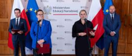 Na tle polskiej i unijnej flagi, na jasnym tle z logami Ministerstwa Edukacji Narodowej, stoi czworo dorosłych. Po lewej stronie od środka, kobieta w okularach, niebieskiej marynarce i czarnych spodniach stoi z mikrofonem przy ustach, trzymając czerwoną teczkę w lewej ręce. Obok niej stoi mężczyzna w granatowym garniturze z krawatem, trzymający czerwoną teczkę.