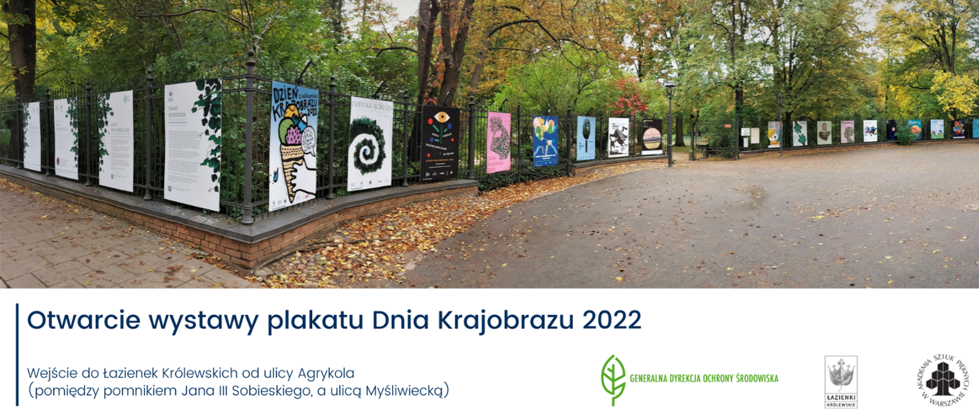 Na płocie widoczne plakaty. Za płotem zielone krzewy i drzewa.