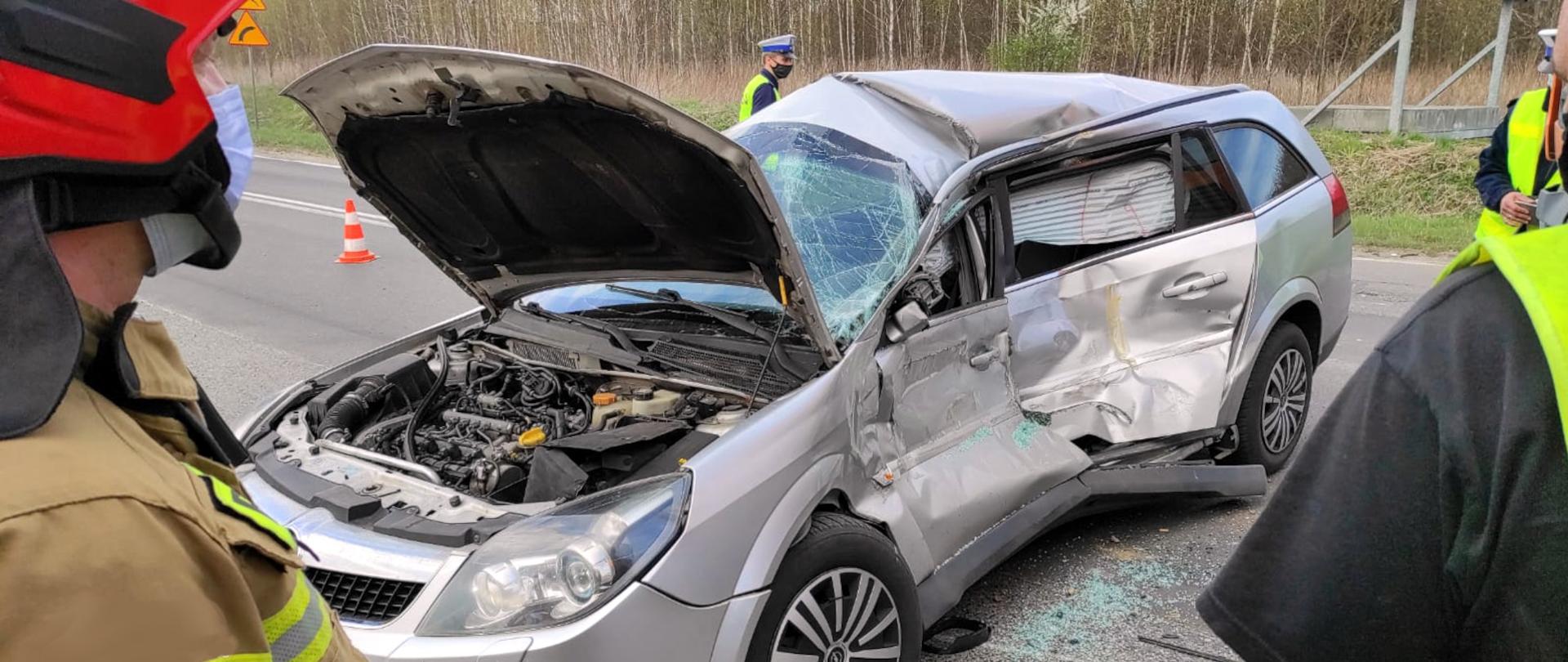 Zdjęcie przedstawia wrak rozbitego samochodu osobowego. Obok stoi 3 policjantów i strażak.