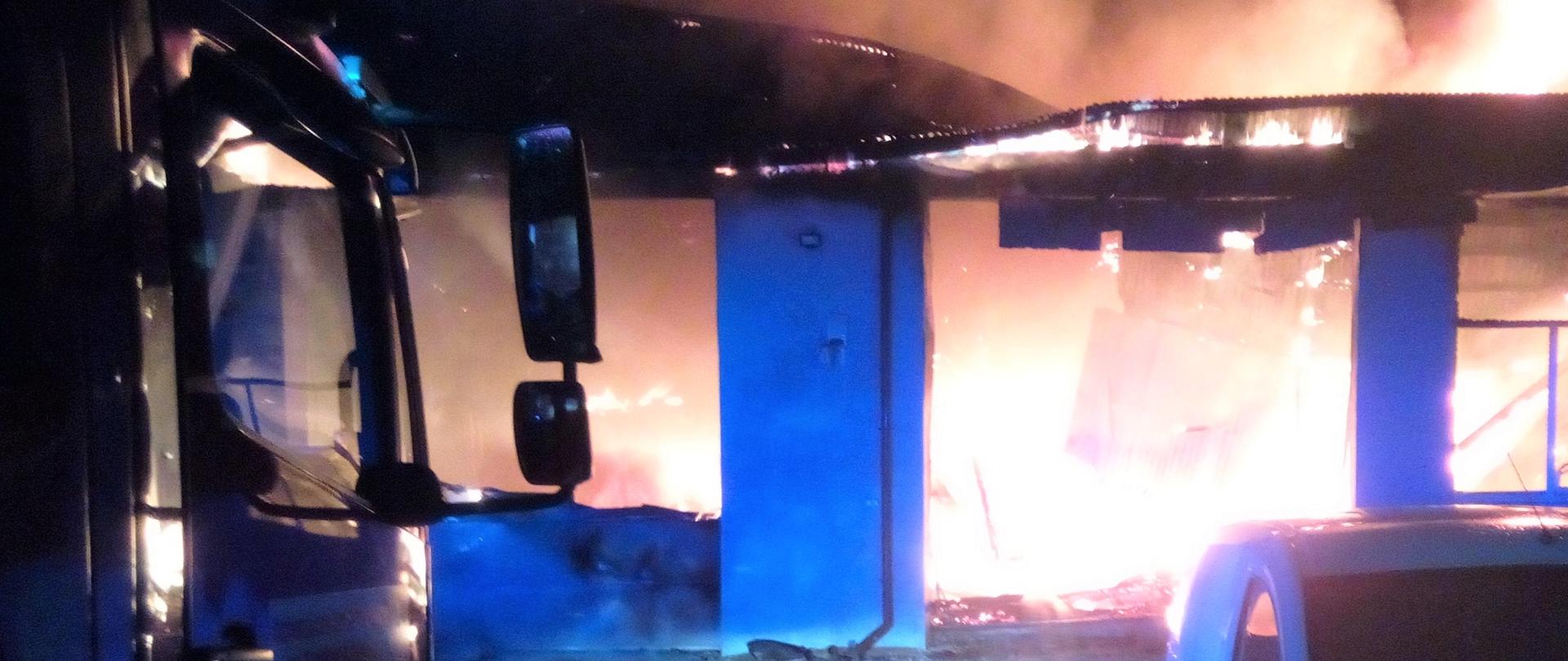 Na zdjęciu przedstawiono rozwinięty pożar w budynku warsztatowym. Po lewej fragment samochodu pożarniczego biorącego udział w działaniach.
