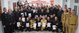 Na zdjęciu w sali konferencyjnej Komendy Powiatowej Państwowej Straży Pożarnej widoczni strażacy Państwowej Straży Pożarnej i druhowie kończący szkolenie podstawowe strażaków ratowników Ochotniczych Straży Pożarnych ubrani w ubrania koszarowe w czarnym i piaskowym kolorze podczas uroczystego zakończenia szkolenia.