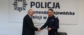 dwaj mężczyźni stoją na tle białej ściany z napisem Policja Komenda Wojewódzka Policji w Łodzi mężczyzna z lewej strony zdjęcia ubrany jest garnitur, mężczyzna z prawej strony zdjęcia ubrany jest w mężczyźni patrzą na siebie z uśmiechem i podają sobie dłoni każdy z nich trzyma teczkę z dokumentami