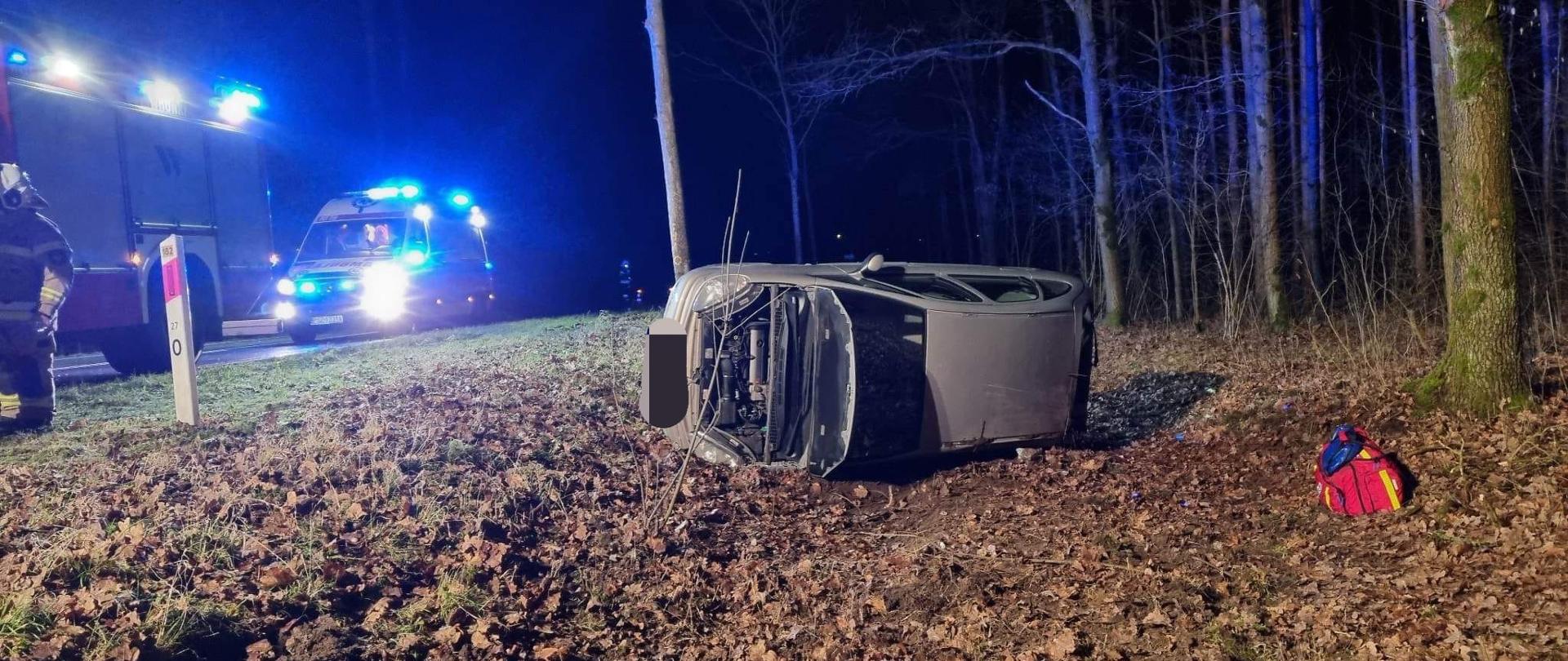 Widać samochód leżący na lewym boku w rowie, przy drzewie, otwarta maska jest noc, po lewej karetka pogotowia, po prawej drzewa w lesie