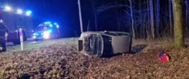 Widać samochód leżący na lewym boku w rowie, przy drzewie, otwarta maska jest noc, po lewej karetka pogotowia, po prawej drzewa w lesie