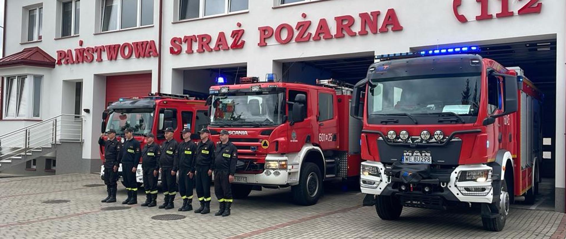 Zdjęcie przedstawia fasadę remizy Państwowej Straży Pożarnej w Przysusze. Budynek jest biało-czerwony, z dużymi drzwiami garażowymi, a nad nimi widnieje napis "PAŃSTWOWA STRAŻ POŻARNA" oraz numer alarmowy "(112)". Przed remizą stoją w rzędzie trzy czerwone wozy strażackie. Dwa z nich to duże samochody gaśnicze, z których jeden ma numer "601-25" i jest marki Scania, a drugi to MAN z rejestracją "WE 8U298" i włączonymi niebieskimi światłami. Trzeci, mniejszy pojazd znajduje się po lewej stronie. Przed wozami, na wybrukowanym placu, stoi szereg strażaków w ciemnych ubraniach koszarowych. Dzień jest pochmurny.