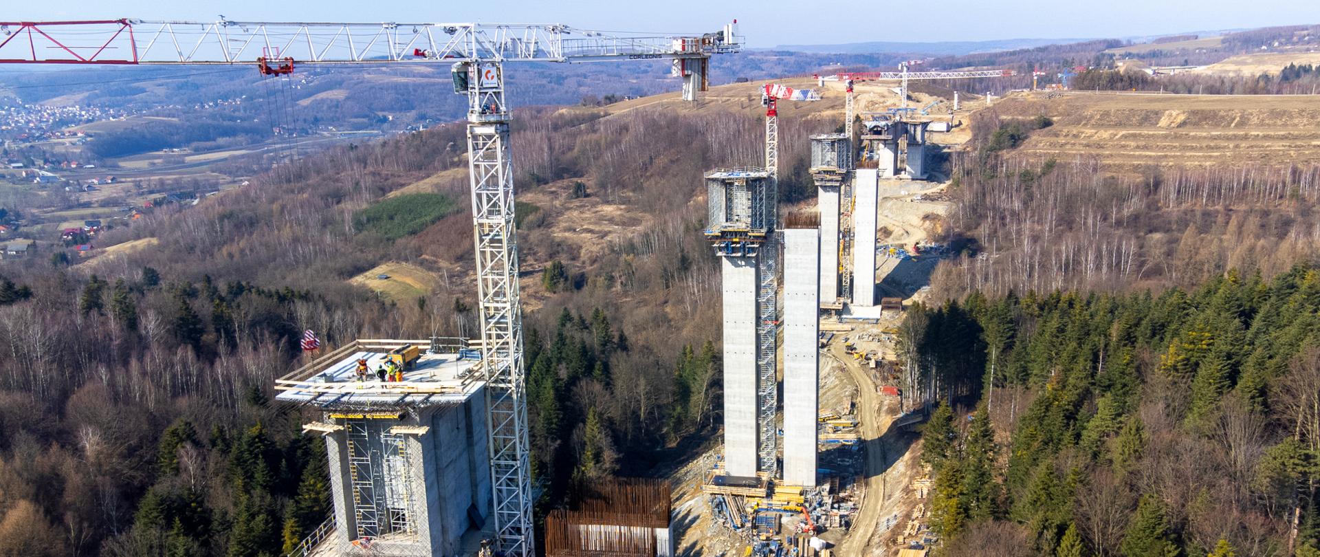 Zdjęcie z lotu ptaka pokazuje budowę dużego estakadowego mostu w terenie górzystym i leśnym. Widać kilka bardzo wysokich, betonowych filarów ustawionych wzdłuż przyszłej trasy drogi, część z nich jest jeszcze w trakcie budowy. Obok filarów stoją duże żurawie wieżowe, a u podnóża konstrukcji znajdują się maszyny i zaplecze budowy. Otoczenie stanowią lasy i pagórkowaty krajobraz.