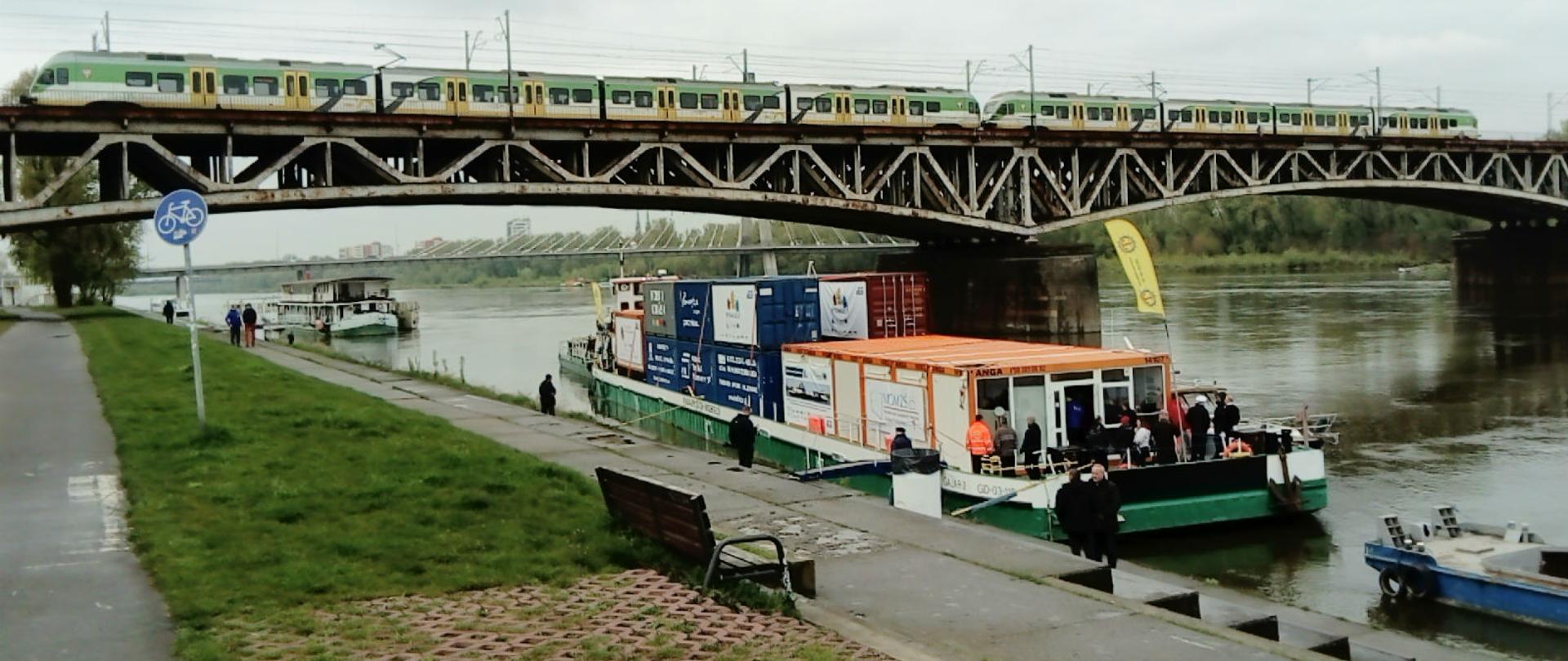 Na zdjęciu pokazana jest barka z kontenerami, która jest zacumowana przy brzegu Wisły w Warszawie