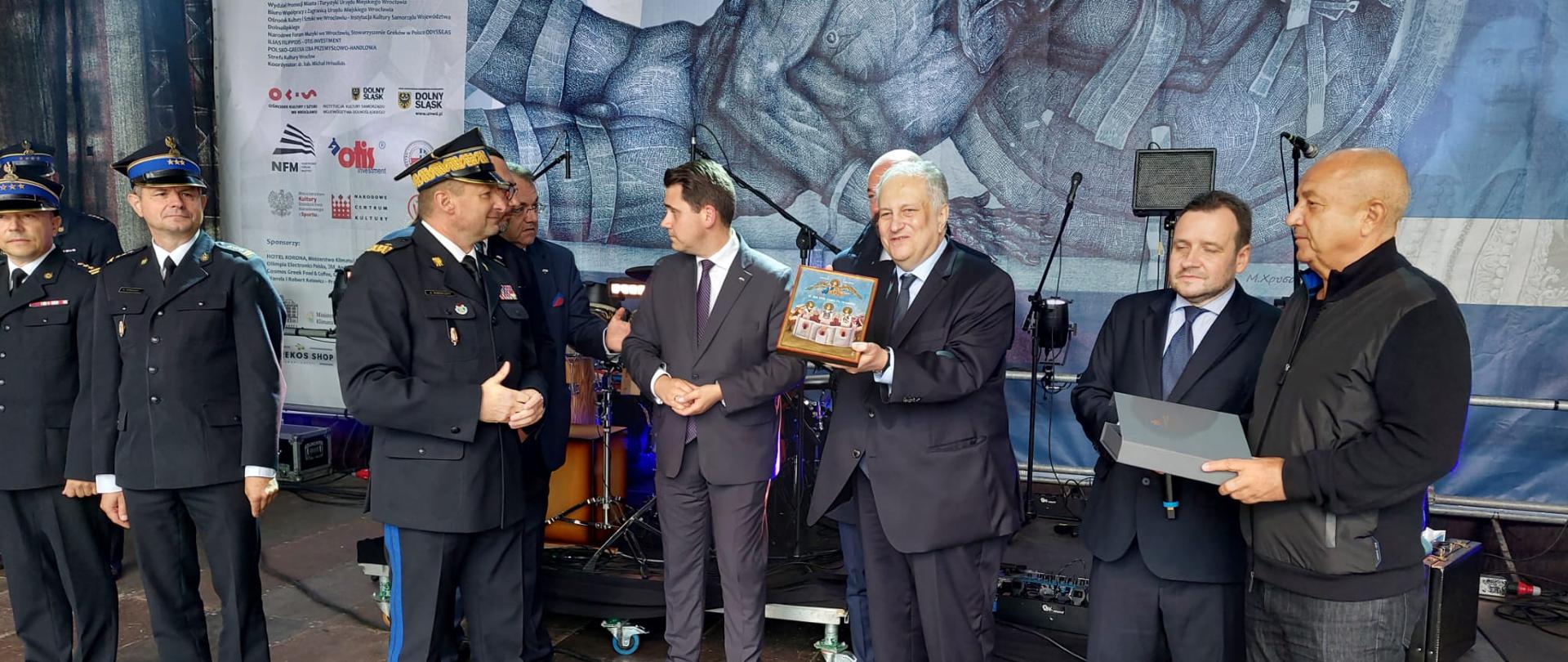 Zastępca KG PSP i inni mężczyźni na scenie podczas podziękowań za misję polskich strażaków, Ambasador Grecji prezentuje w dłoniach ikonę, którą otrzyma Zastępca KG PSP
