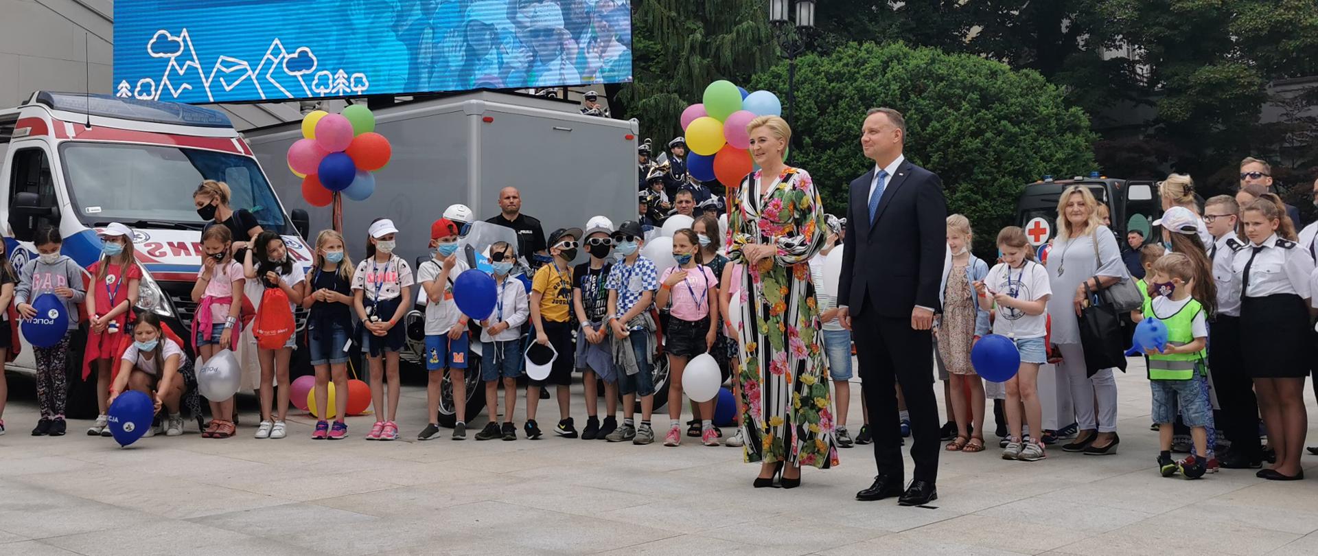 Prezydent RP z małżonką stoją na dziedzińcu ogrodu prezydenckiego w tle grupa dzieci i telebim z hasłem Bezpieczne Wakacje z Parą Prezydencką
