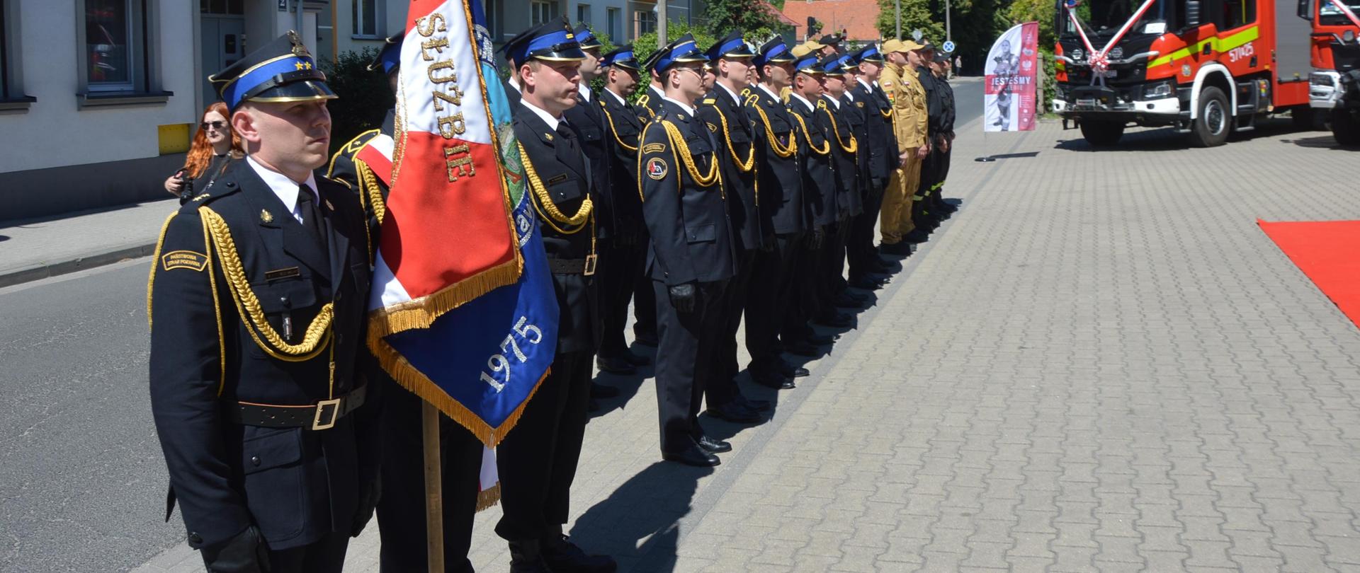 Dzień Strażaka 2024 Plac przed komendą. W dwuszeregu ustawiony jest pododdział funkcjonariuszy KP PSP w Rawiczu. Na pierwszym planie znajduje się poczet sztandarowy. W tle dwa wozy bojowe. 