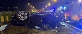 Zdjęcie przedstawia miejsce wypadku drogowego, które miało miejsce nocą na autostradzie. Na pierwszym planie widać samochód osobowy leżący na dachu, częściowo znajduje się na poboczu trawy, a częściowo na asfalcie. Pojazd jest uszkodzony – zniekształcony dach, wyłamane części karoserii. W tle widać kilka pojazdów służb ratunkowych – w tym wóz straży pożarnej z włączonymi światłami ostrzegawczymi. Na miejscu pracują strażacy w hełmach. Wzdłuż drogi znajdują się barierki ochronne oraz oświetlenie uliczne.