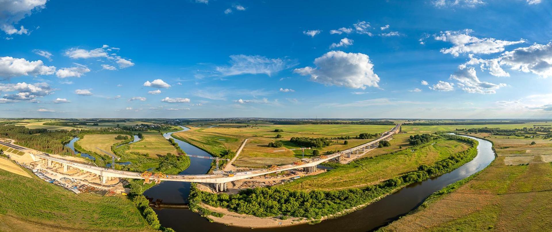 Widok z lotu ptaka na wielką plac budowy mostu nad meandrującą rzeką. Rzeka wygina się przez krajobraz, zielone pola i skupiska drzew po obu stronach. Na placu budowy widać wiele żurawi, kilka częściowo ukończonych odcinków mostu z widocznymi rusztowaniami oraz liczne pojazdy budowlane. Niebo jest bezchmurne, co sugeruje dobre warunki pogodowe do prac budowlanych.