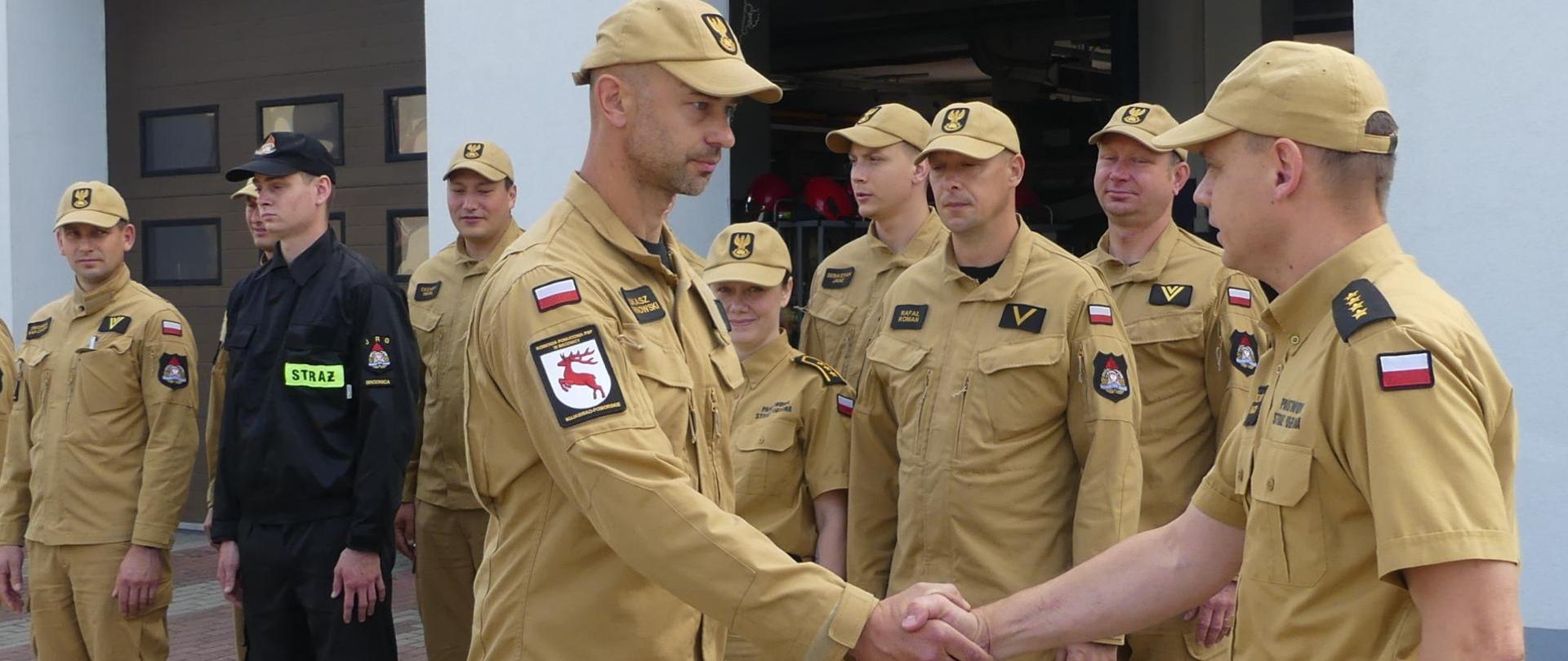 Zdjęcie przedstawia Komendanta Powiatowego PSP w Brodnicy ściskającego dłoń ogn. Łukasza Sarnowskiego podczas gratulacji z okazji jubileuszu 25 lat pracy i służby. Za nimi stoją funkcjonariusze Komendy. Za nimi budynek JRG w Brodnicy.