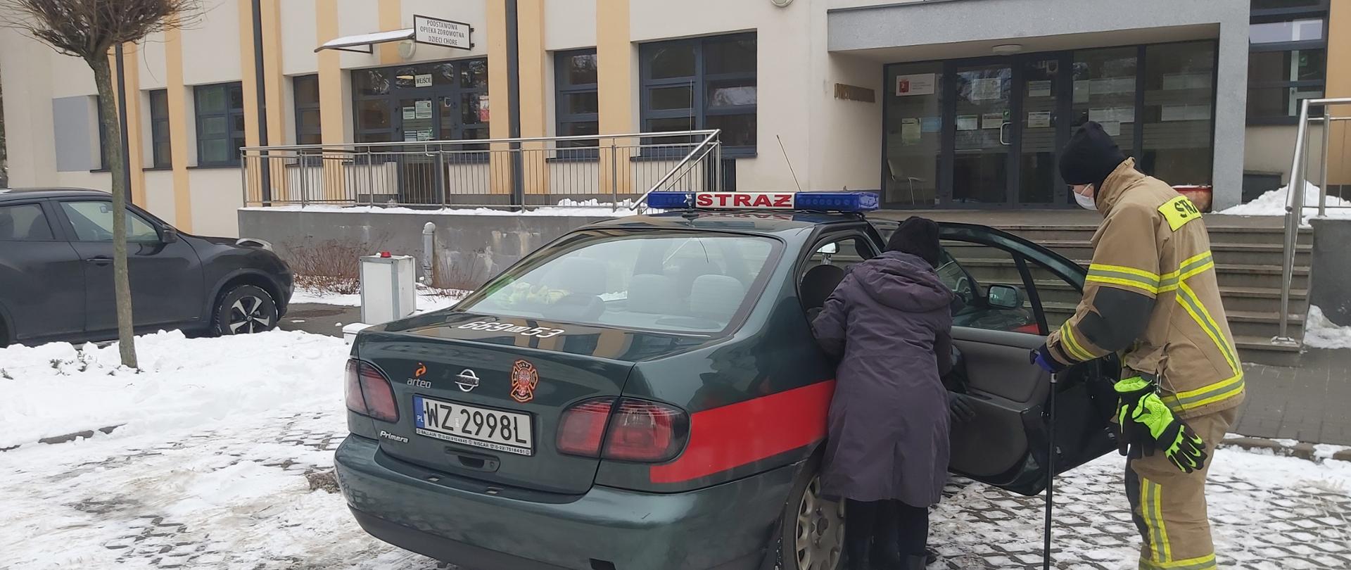 Na zdjęciu widać wsiadająca starszą kobietę wsiadającą do samochodu strażackiego w asyście strażaka