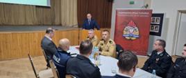 Strażacy ubrani w granatowe mundury galowe, oraz pisakowe mundury siedzą przy stolikach, w tyle rolap z logiem Państwowej Straży Pożarnej na tylnej ścianie kotary oraz ekran z wyświetloną prezentacją. Przy mównicy stoi starosta ubrany w garnitur, przed sobą ma mikrofon