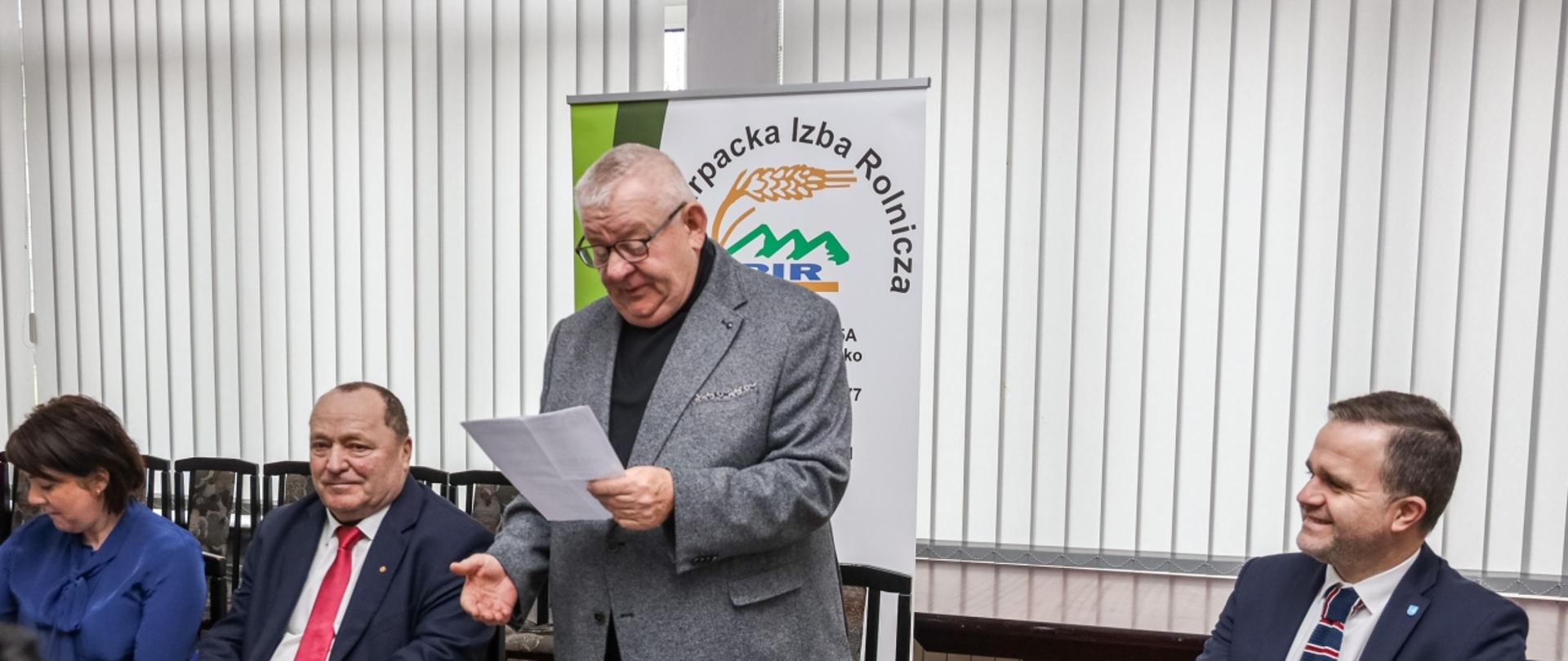 Czterech mężczyzn i jedna kobieta. Ubrani są na elegancko w garnitury. Siedzą przy czarnym stole na którym leżą dokumenty oraz butelki z wodą. Jeden z mężczyzn stoi, trzymając w dłoniach kartkę i czyta. W tle stoi baner z logiem instytucji. Za banerem jasne rolety.