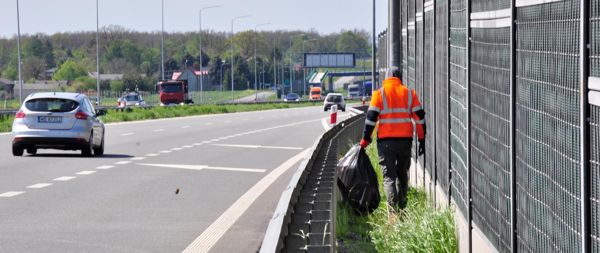 Zdjęcie przedstawia osobę ubraną w pomarańczową kamizelkę odblaskową, która porusza się wzdłuż pobocza autostrady, zbierając śmieci. Osoba ta trzyma duży czarny worek na śmieci i idzie obok metalowego ogrodzenia dźwiękochłonnego. Na drodze w tle widoczne są pojazdy, w tym srebrny samochód osobowy oraz czerwona ciężarówka. Po lewej stronie zdjęcia znajduje się pas zieleni oddzielający jezdnię od pobocza. W tle widoczne są drzewa oraz budynki, a dzień jest pogodny, z lekkim zachmurzeniem.