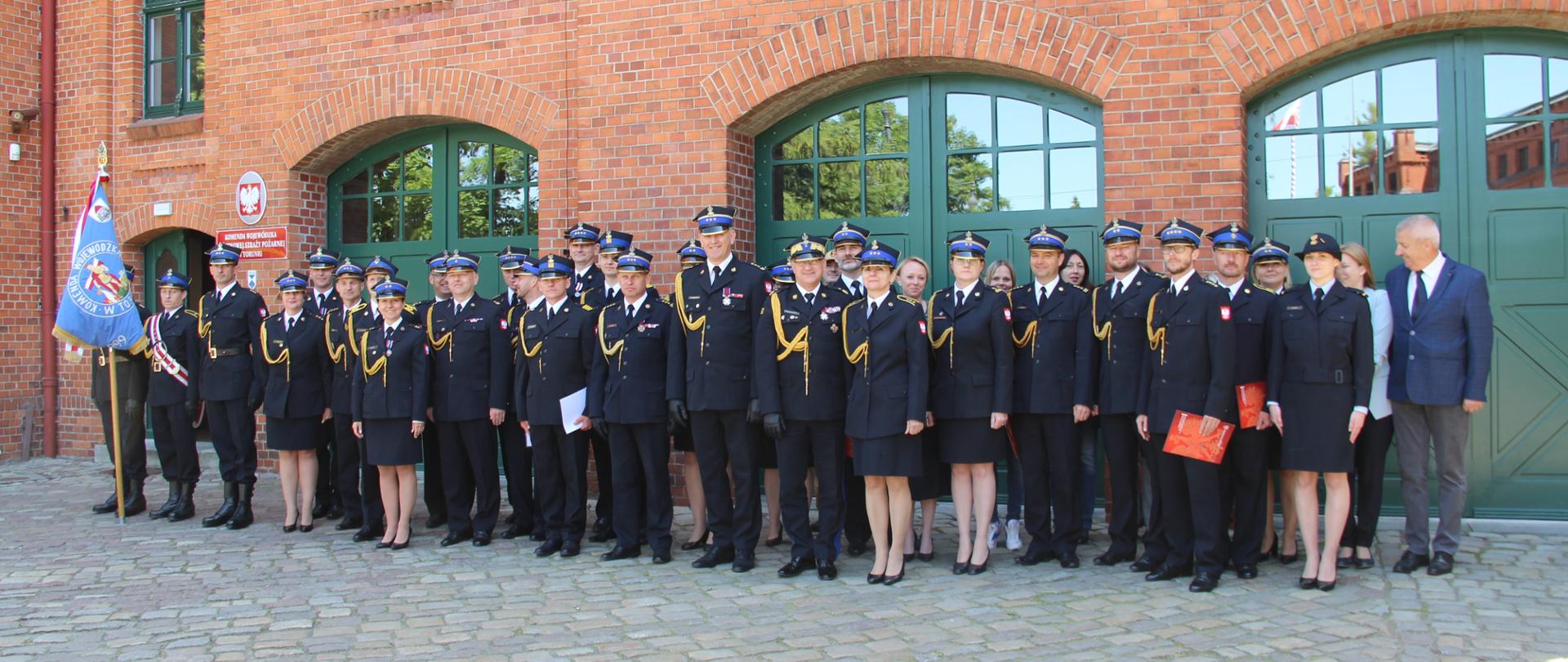 Zdjęcie grupowe pracowników KW PSP Toruń. Za nimi budynek siedziby. Z lewej strony poczet sztandarowy.