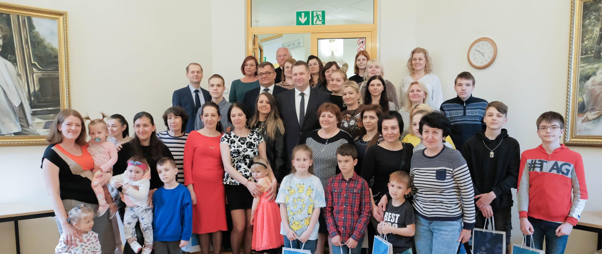 Minister Przemysław czarnek w grupie dzieci i ich matek. Dzieci trzymają w ręku torby z prezentami. W tle zegar i obrazy 