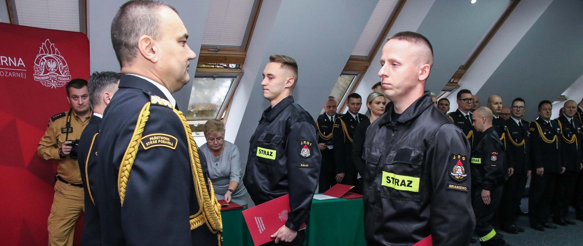 Sala konferencyjna. Przed komendantem wojewódzkim dwóch strażaków przyjmuje ślubowanie. W tle strażacy w mundurach wyjściowych.
