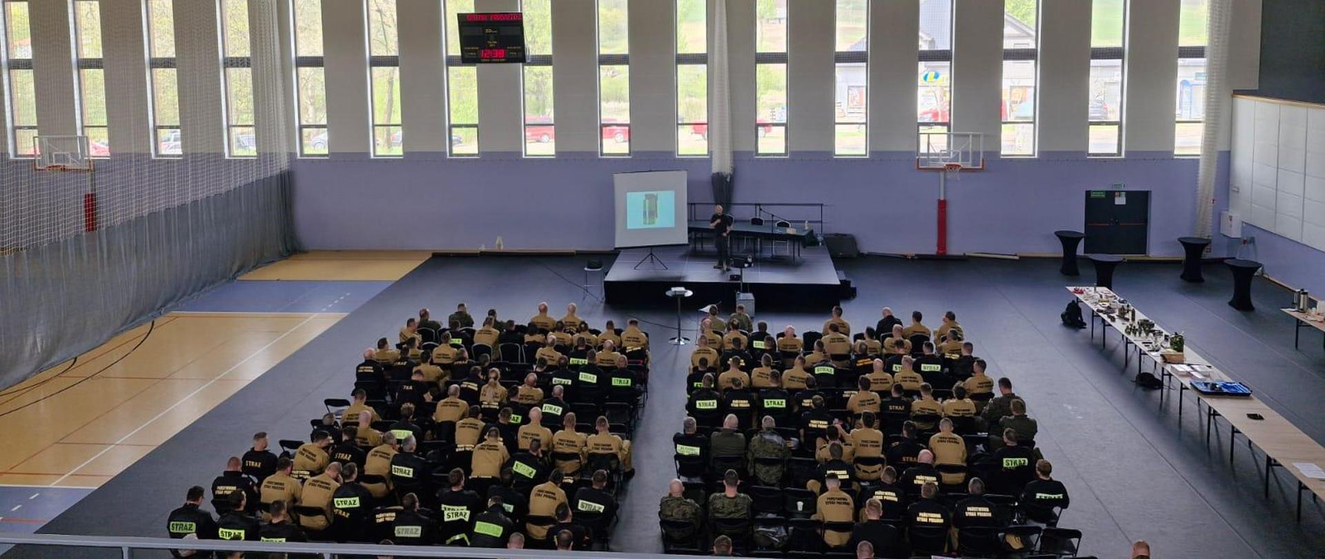 Strażacy w mundurach kolory musztardowego oraz czarnego siedzą na krzesłach przed nimi na podeście stoi mężczyzna który prowadzi szkolenie na ekranie multi medialnym wyświetlana jest prezentacja obok na stołach rozłożony jest poczęstunek.