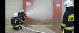 Trzech mężczyzn w ubraniach bojowych strażackich, w białych hełmach polewają drzwi budynku wodą. Jest to element ćwiczenia.