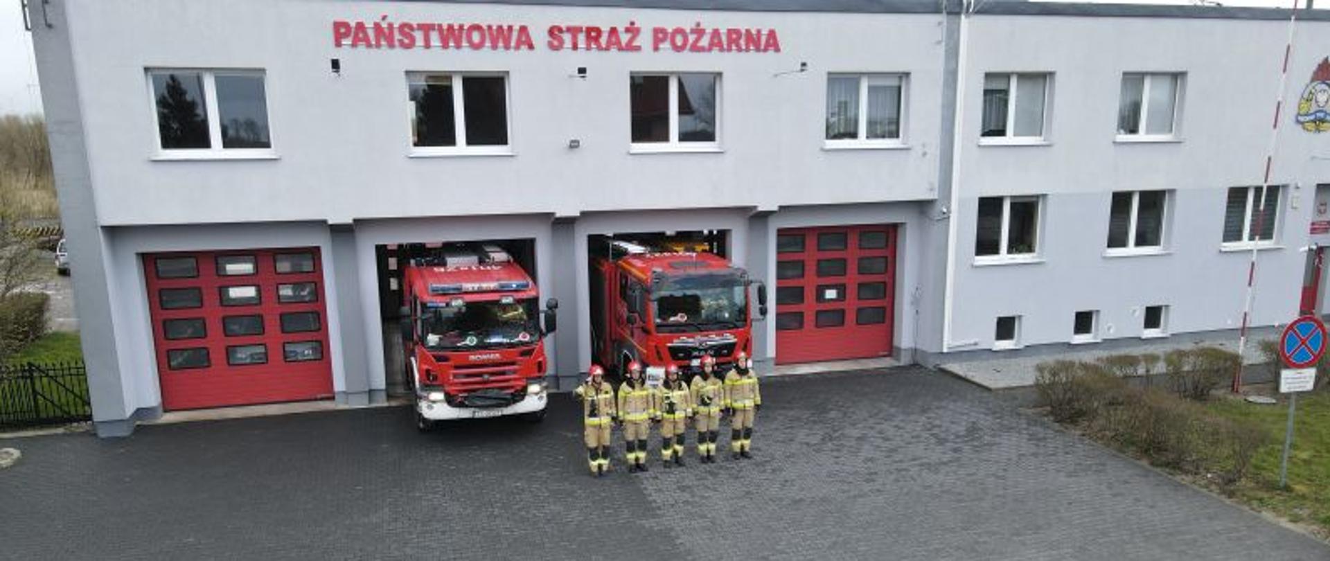 Zdjęcie wykonane w dzień. Przed budynkiem Komendy stoją dwa wozy strażackie. Przed wozami zmiana oddająca hold ofiarom katastrofy Smoleńskiej. Strażacy ubrani w piaskowe mundury i czerwone hełmy.