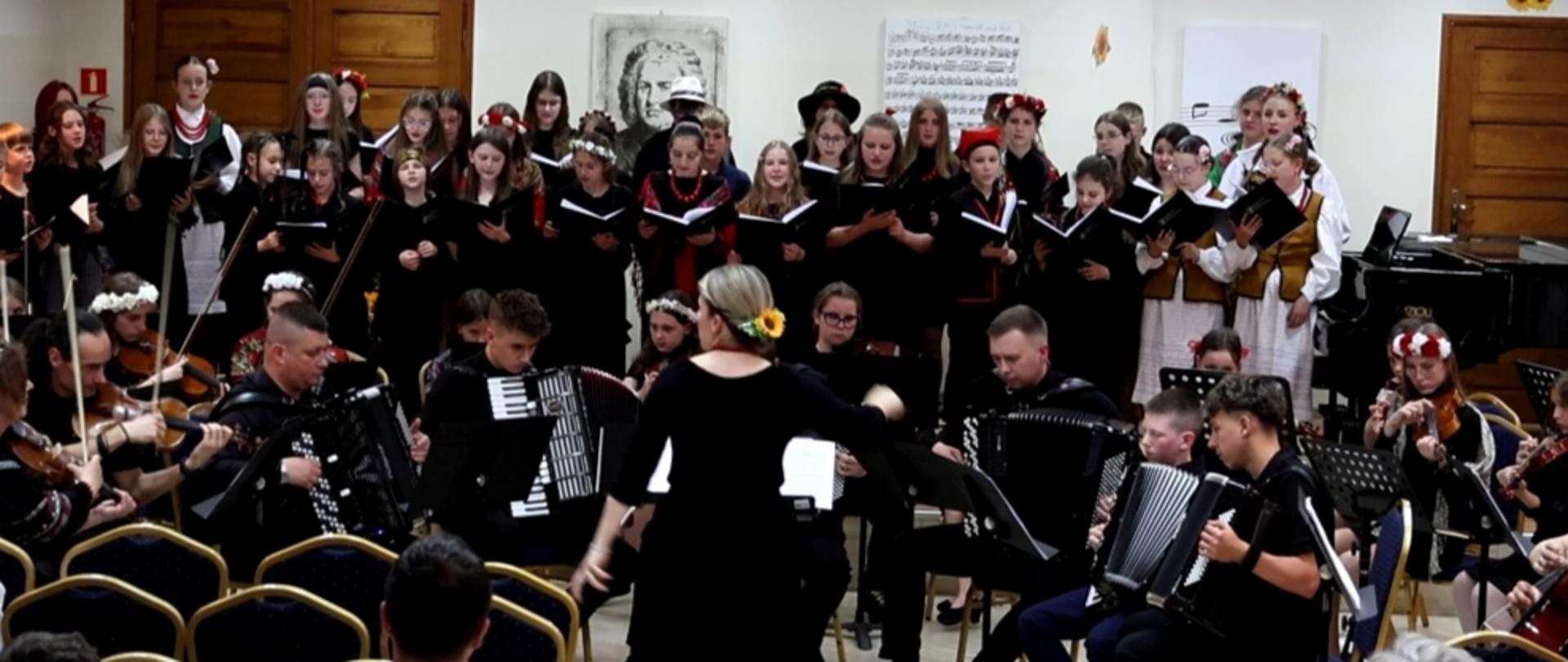 Na zdjęciu widać występ muzyczny w sali koncertowej. Na pierwszym planie dyrygentka prowadzi orkiestrę złożoną głównie z akordeonistów i skrzypków. W tle stoi duży chór dzieci i młodzieży ubrany w ciemne stroje, część osób ma na głowach tradycyjne wianki. W sali ustawiono krzesła dla publiczności, a za wykonawcami znajdują się obrazy i fortepian. Atmosfera wydarzenia jest uroczysta i artystyczna.