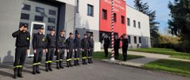 Strażacy stoją w szeregu przy maszcie flagowym. Poczet flagowy i dowódca salutują. Za strażakami budynek komendy w kolorach szaro - czerwonych. W prawej części zdjęcia skoszony trawnik i drzewo iglaste. 