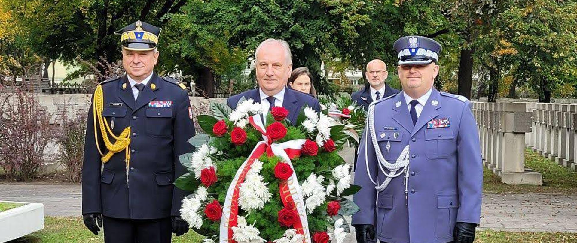 Wojewoda pomorski trzyma wieniec funkcjonariusze podlegli Ministerstwu Spraw Wewnętrznych i Administracji stoją po obu stornach mężczyzny trzymającego wieniec.