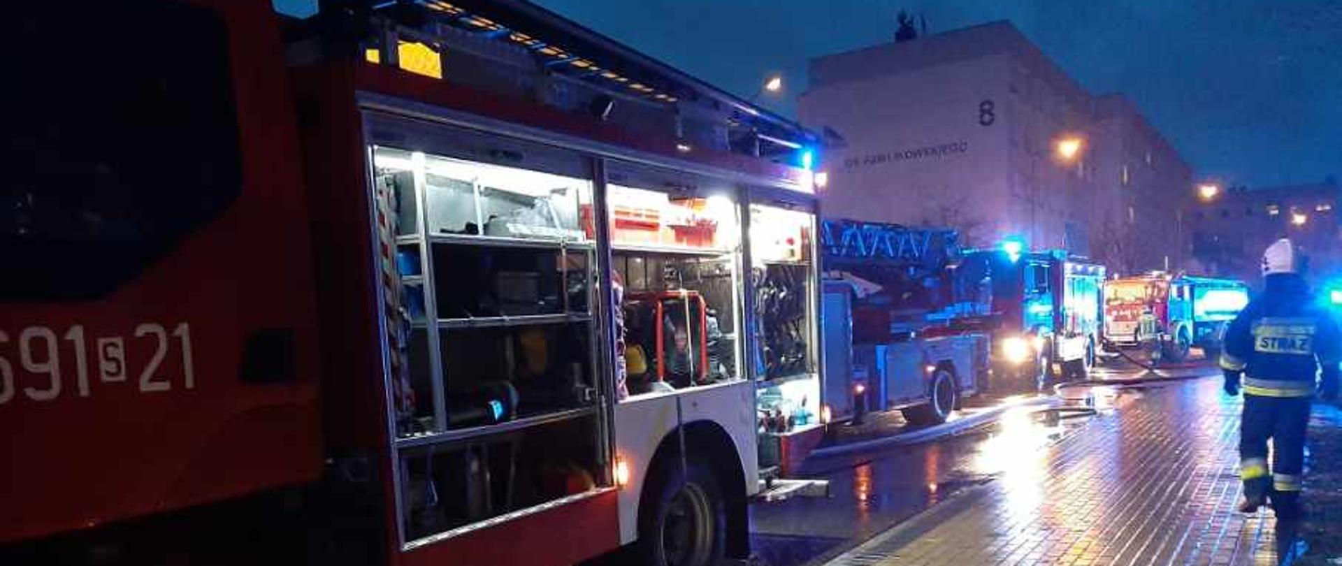 Samochody strażackie z włączonymi sygnałami świetlnymi, widoczne od ich lewego boku. Pierwszy ma otwarte skrytki sprzętowe. Po prawej stronie zdjęcia strażak w białym hełmie. W tle bloki mieszkalne. 