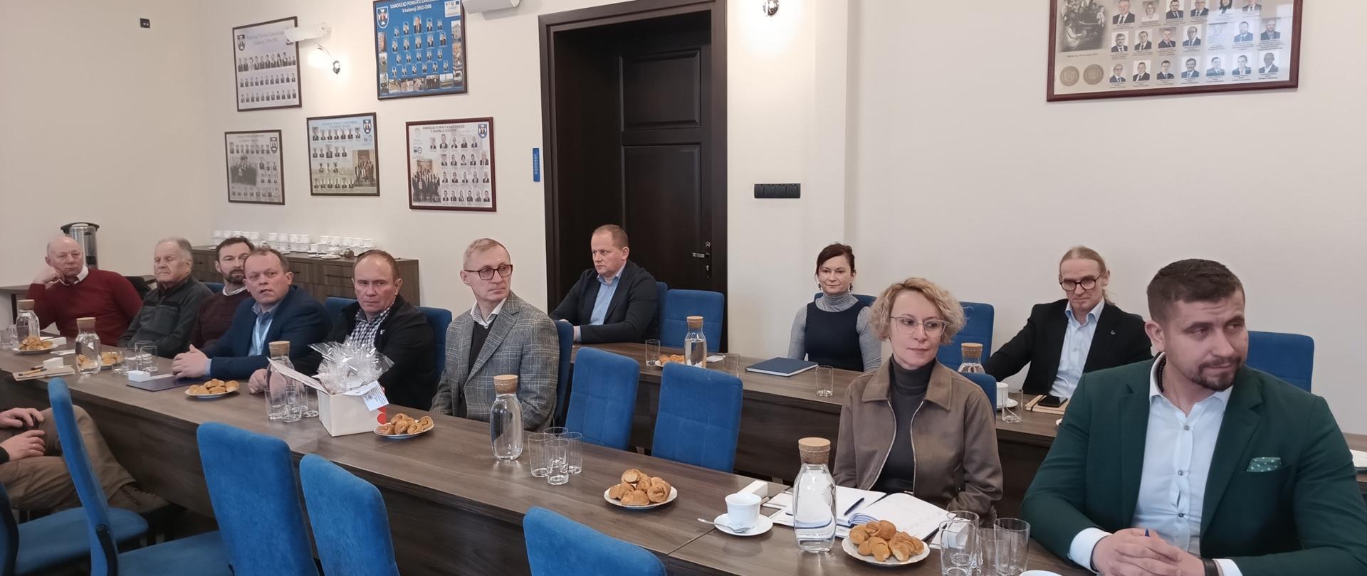 Grupa ludzi siedząca na niebieskich krzesłach przy brązowym stole. Dwie kobiety i dziewięć mężczyzn. Mężczyzna na pierwszym planie ubrany jest elegancko w ciemno zieloną marynarkę. Kobieta obok niego ubrana w brązową kurtkę i ciemny golf. Znajdują się w jasnym pomieszczeniu z brązowymi drzwiami. Na ścianach wiszą obrazy ze zdjęciami. Na stołach przed nimi znajdują się dzbanki z wodą ,szklanki i talerzyki z rogalikami