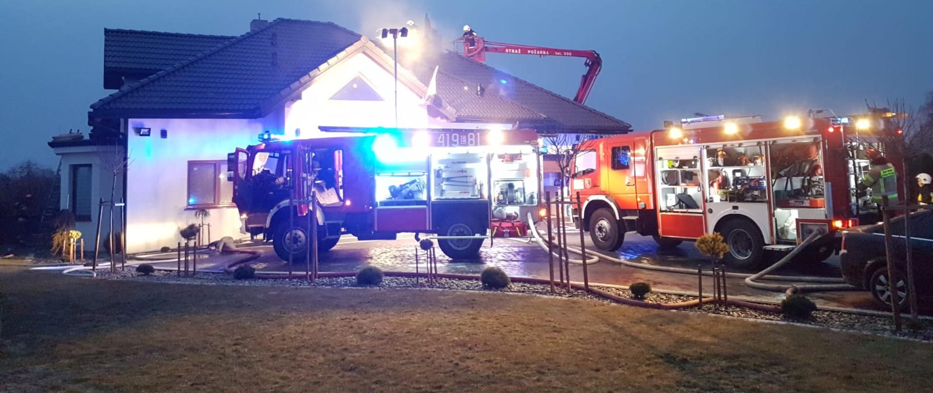 Budynek mieszkalny jednorodzinny w tle. Na pierwszym planie dwa wozy pożarnicze. Trwa akcja gaśnicza podnośnikiem hydraulicznym na dachu budynku skąd wydobywają się kłęby dymu.