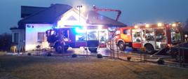 Budynek mieszkalny jednorodzinny w tle. Na pierwszym planie dwa wozy pożarnicze. Trwa akcja gaśnicza podnośnikiem hydraulicznym na dachu budynku skąd wydobywają się kłęby dymu.