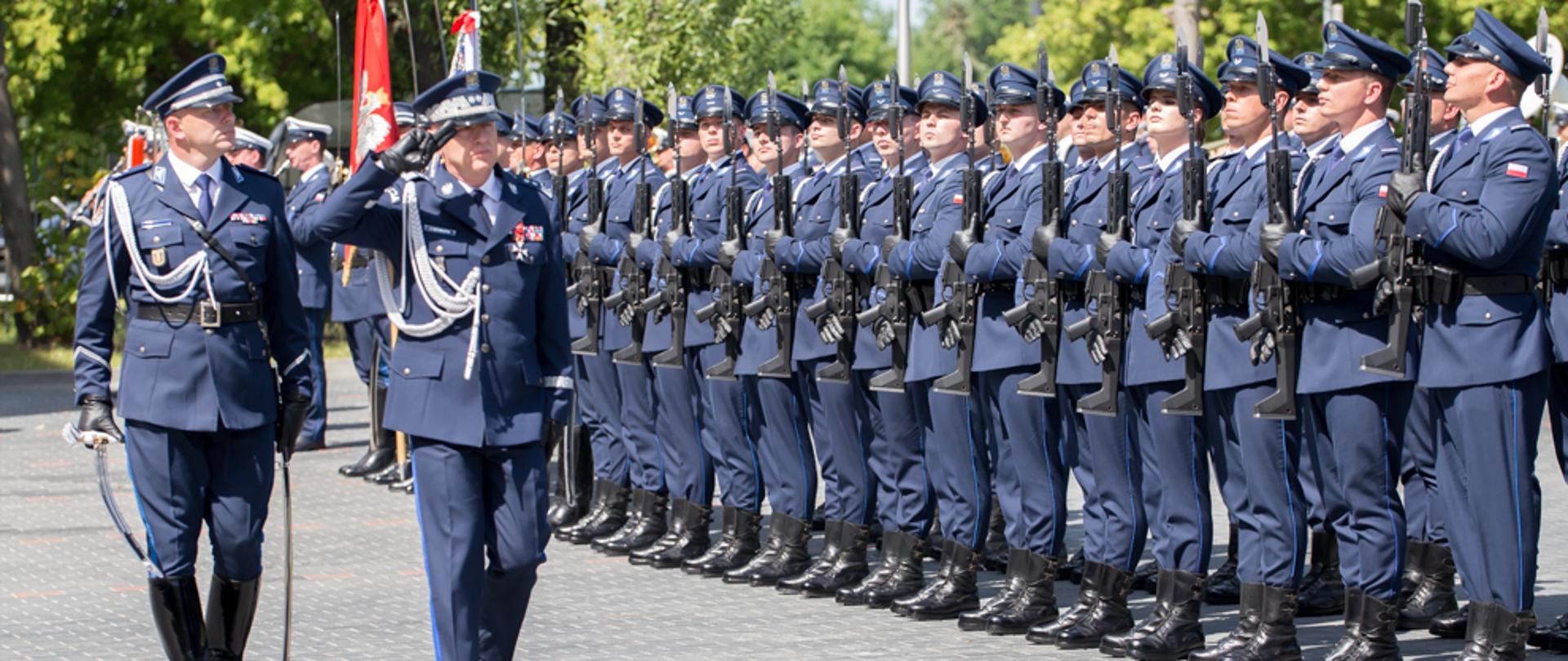 Przegląd pododdziałów podczas obchodów Święta Policji. Komendant Główny Policji dokonuje przeglądu pododdziałów podczas obchodów Święta Policji. Oddaje honor przez salutowanie przed kompanią reprezentacyjną Policji. Za nim idzie w asyście Dowódca Uroczystości, który w prawym ręku trzyma szablę. 