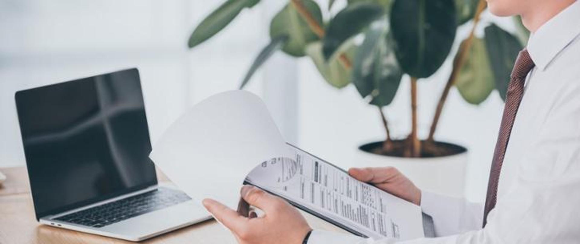 Na zdjęciu widoczny jest mężczyzna trzymający w rękach dokumenty. W tle widoczny jest laptop leżący na biurku. Obok znajduje się kwiat.