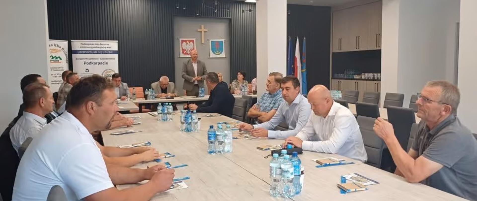 Grupa ludzi siedzi przy stołach konferencyjnych. Jeden mężczyzna stoi i przemawia. 