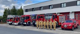 Sześciu strażaków w jasnych mundurach stoi na baczność przed kilkoma wozami strażackimi i czerwonym samochodem terenowym, zaparkowanymi przed nowoczesnym budynkiem.