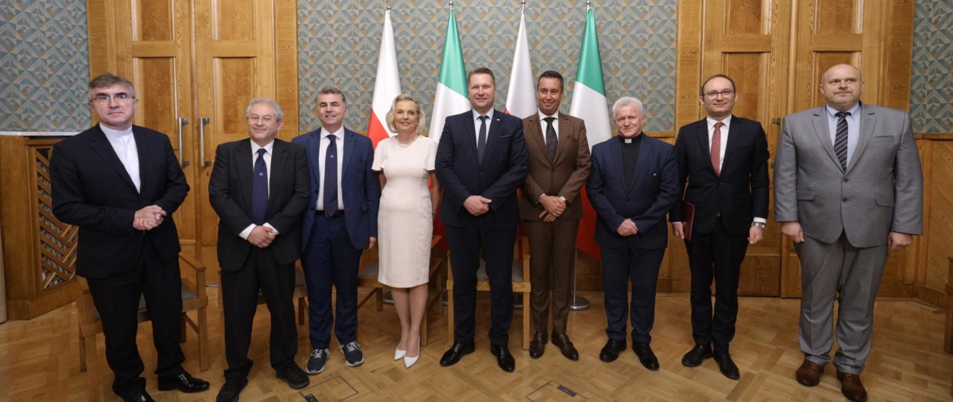 Zdjęcie zbiorowe uczestników spotkania, stoją w rzędzie pod ścianą, za nimi polskie i włoskie flagi.