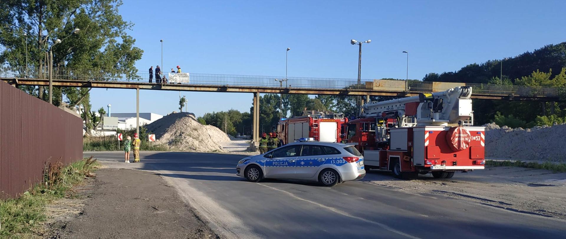 Wozy strażackie stoją po prawej stronie ulicy u góry most/kładka z której próbuje skoczyć mężczyzna, obok niego funkcjonariusze w trakcie rozmowy.