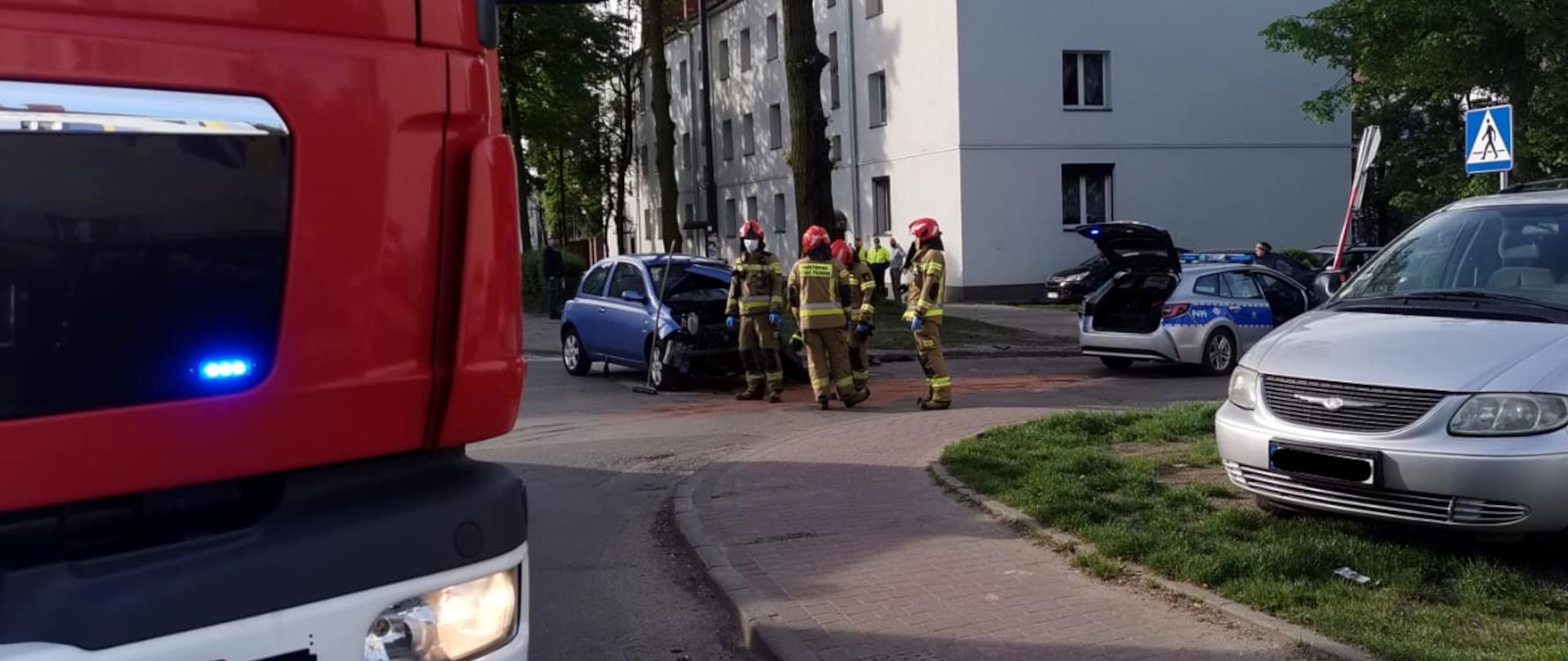 Zdjęcie przedstawia czterech strażaków likwidujących skutki kolizji. Obok nich stoją rozbite samochody uczestniczące w kolizji oraz policyjny radiowóz.